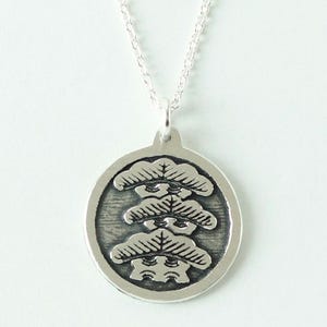 Pode incluir: Colar de prata esterlina com um pendente redondo com um design de árvore estilizado e detalhado. O pendente tem 19 mm de diâmetro e está pendurado numa corrente fina. O texto "Sterling Silver" é visível. Fundo branco.