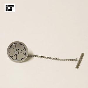 Puede incluir: Un pasador de corbata plateado con un diseño floral. El pasador presenta una cara circular con un motivo de flor, conectado por una cadena a un cierre en forma de T. El diseño es simple y elegante, adecuado para vestimenta formal.