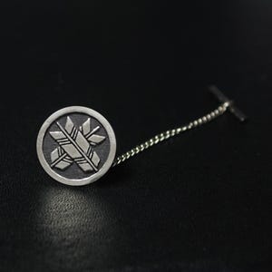 Puede incluir: Pin de corbata plateado con una cabeza circular con un diseño estilizado de flechas cruzadas y formas geométricas. El pin tiene una cadena y un cierre en T. El fondo es oscuro.