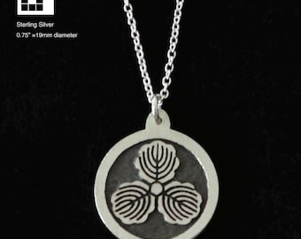 Może przedstawiać: Naszyjnik z wisiorkiem ze srebra próby 925. Wisiorek ma okrągły kształt z kwiatowym emblematem. Wisiorek ma średnicę 19 mm i wisi na delikatnym łańcuszku. Naszyjnik jest pokazany na ciemnym tle.