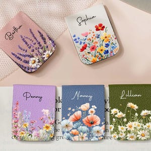 May include: A collection of personalized magnetic bookmarks with floral designs. Each bookmark features a name: Bertha, Sophia, Penny, Nancy, and Lillian. The bookmarks are in various colors, including pink, white, purple, blue, and green.