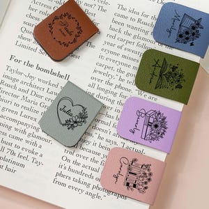 Op de afbeelding: Leren bladwijzers in diverse kleuren, elk met een bloemmotief en een naam. De bladwijzers, in bruin, blauw, groen, paars en roze, liggen op een open boek.
