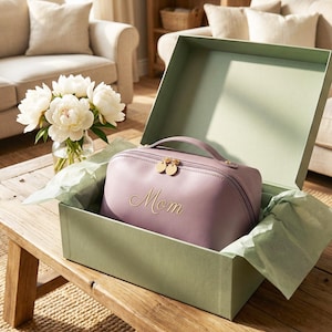 Puede incluir: Un neceser lavanda con la palabra "Mamá" en escritura dorada se presenta dentro de una caja de regalo verde claro. La bolsa tiene un asa y cremalleras doradas. La caja está sobre una mesa de madera, con un ramo de peonías blancas cerca.