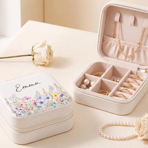 May include: A square white jewelry box with a floral print and the name Emma printed on the lid. The open box features a beige interior with compartments for rings and a necklace holder. A pearl bracelet and two dried roses are placed nearby.