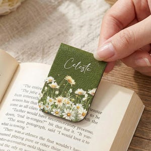 Könnte beinhalten: Grüner Lesezeichen mit dem Namen "Celeste" in Schreibschrift. Das Lesezeichen zeigt ein Blumendesign aus weißen Gänseblümchen mit gelben Zentren. Das Lesezeichen liegt auf den Seiten eines Buches.