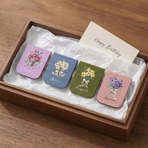 Puede incluir: Una caja de madera contiene cuatro marcapáginas con temática floral en morado, azul, verde y rosa. Cada marcapáginas presenta un ramo de flores diferente y un nombre. También hay una tarjeta con "Happy Birthday".