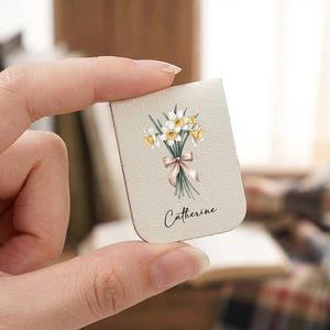 Puede incluir: Marcapáginas beige con una ilustración de acuarela de narcisos, atados con una cinta rosa. El nombre "Catherine" está escrito debajo de las flores. El marcapáginas se sostiene en una mano.