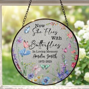 May include: A circular glass suncatcher hanging by a black chain, featuring a textured surface with a floral and butterfly print. The text reads: Now She Flies With Butterflies In Loving Memory Amelia Smith 1972-2025, surrounded by colorful wildflowers.