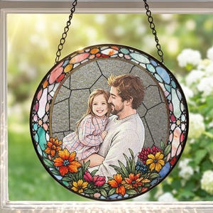 May include: A circular stained glass suncatcher featuring a central portrait of a person holding a young child. The border is decorated with colourful floral patterns in orange, yellow, pink, and blue, suspended by a black metal chain.