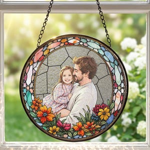May include: Circular stained glass window art depicting a father and child. The image is framed by colourful floral and geometric patterns. The window is suspended by a black chain, set against a bright window.