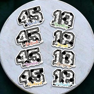 Pode incluir: Uma coleção de autocolantes com tema de futebol com os números 45 e 18 em preto e branco. Cada autocolante inclui um nome em fonte cursiva, como Charles, Bradley, Patrick e outros. Os autocolantes estão dispostos numa superfície redonda de cor clara.