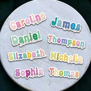May include: A selection of colourful name stickers on a white wooden surface. Each sticker displays a first and last name in a playful, retro font. Names include Caroline Taylor, James Smith, Daniel Anderson, and others.