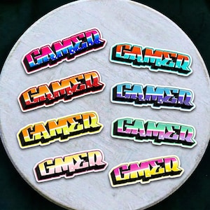 Puede incluir: Una colección de pegatinas "GAMER" coloridas en varios diseños. Cada pegatina presenta la palabra "GAMER" en una fuente audaz de estilo grafiti con un contorno blanco. Las pegatinas están dispuestas sobre una superficie circular gris claro.