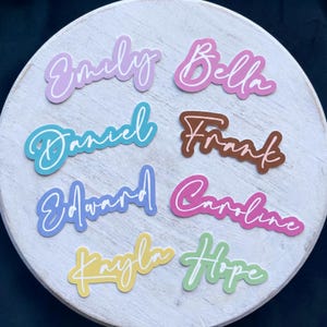 Puede incluir: Una colección de nombres recortados en colores vibrantes, cada uno con una fuente de escritura única y un contorno blanco. Los nombres incluyen Emily, Bella, Daniel, Frank, Edward, Caroline, Kayla y Hope. Dispuestos sobre una superficie de madera redonda y blanca.