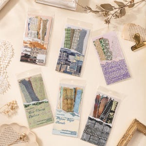 Peut inclure: Six emballages rectangulaires en plastique transparent contenant des bandes de papier décoratif assorties. Chaque paquet présente un thème unique avec des motifs, textures et couleurs variés, incluant des carreaux, des fleurs et des pellicules, avec des étiquettes textuelles imprimées.