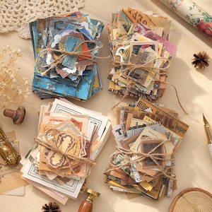 May include: Four stacks of assorted paper ephemera tied with brown twine. The stacks feature various designs including blue landscape art, vintage floral patterns, beige document-style prints, and sepia-toned newspaper clippings with text.