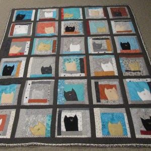 May include: A rectangular quilt featuring a grid of twenty-five square blocks. Each block displays a stylised cat silhouette in black, white, or tan, set against various colourful fabric backgrounds including blue, orange, and grey patterns.