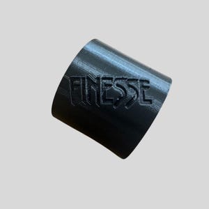 Op de afbeelding: Een zwart, cilindrisch object met het woord "FINESSE" in reliëf op het oppervlak. Het object is licht gekanteld, waardoor de tekst en de gebogen vorm zichtbaar zijn. De achtergrond is effen lichtgrijs.