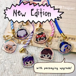May include: A selection of clear acrylic charms with cartoon character illustrations. Each charm showcases a different character design, attached to colourful braided cords with gold-coloured clasps. The text "New Edition" and "with packaging upgrade!" are visible.
