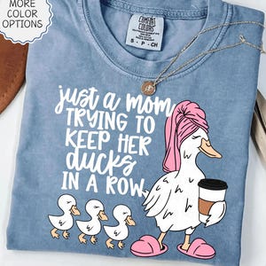 Op de afbeelding: Lichtblauw Comfort Colors T-shirt met de tekst "just a mom trying to keep her ducks in a row". De afbeelding toont een cartoon eend met een roze handdoek en slippers, met een kopje koffie, met drie eendjes.