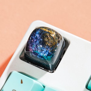 May include: A custom keyboard keycap with a galaxy design. The keycap is a square shape with a clear resin top, showcasing a swirling mix of blue, gold, and purple colors. The keyboard is white with light blue keys.