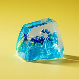 Pode incluir: Uma tecla de resina transparente com um design de escorpião azul e branco. A tecla tem um padrão de ondas azuis e verdes nas laterais. O escorpião é detalhado com listras azuis e brancas, sobre um fundo amarelo.