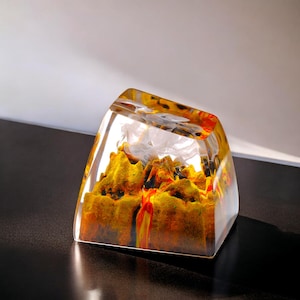 Op de afbeelding: Een heldere, rechthoekige hars papervanger met een berglandschap ontwerp. De binnenkant heeft gele, oranje en rode tinten, waardoor een levendig, artistiek effect ontstaat. De papervanger staat op een donkere ondergrond.