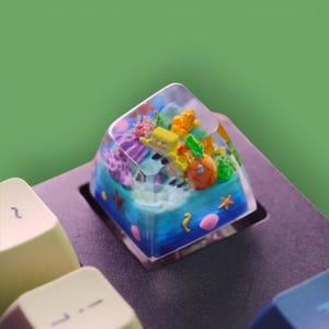 May include: A custom keyboard keycap featuring a miniature underwater scene. The keycap is clear with a blue and turquoise base, showcasing a cartoon character, a pineapple, and coral. The keycap is square and sits on a black keyboard.