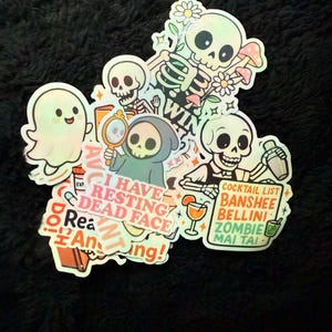 Op de afbeelding: Een verzameling holografische stickers met griezelige skelet- en spookontwerpen. Sommige stickers bevatten tekst zoals "I Have Resting Dead Face" en "Cocktail List". De stickers liggen op een donkere, getextureerde ondergrond.