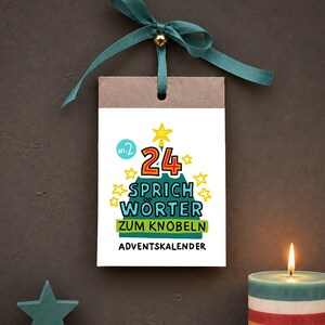 Könnte beinhalten: Ein hängender rechteckiger Adventskalender mit einem blaugrünen Band und einer goldenen Perle. Die weiße Karte zeigt eine grüne Baumgrafik, gelbe Sterne und den Text 24 SPRICH WÖRTER ZUM KNOBELN ADVENTSKALENDER in schwarzer, roter und blauer Schrift.