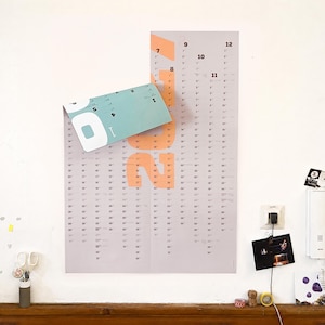 May include: A wall-mounted vertical calendar featuring a light gray background with large peach-colored numbers for the year 2022. The top left corner is folded over, revealing a teal-colored section. The calendar displays months and dates in black text.