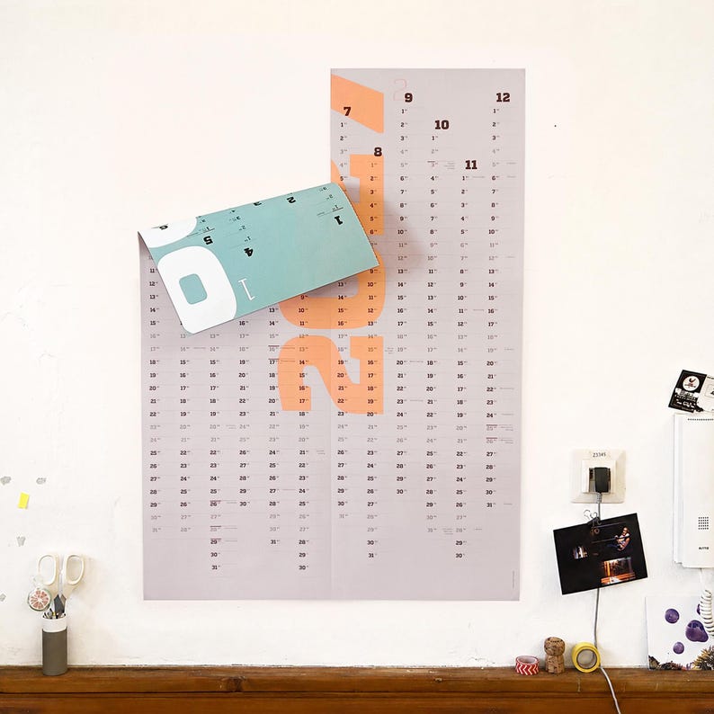 May include: A wall-mounted vertical calendar featuring a light gray background with large peach-colored numbers for the year 2022. The top left corner is folded over, revealing a teal-colored section. The calendar displays months and dates in black text.
