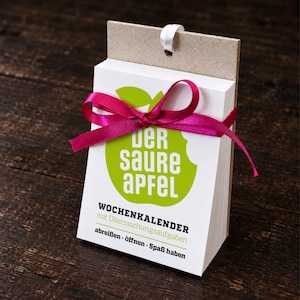 Könnte beinhalten: Ein weißer Abreiß-Wochenkalender mit einer grünen Apfelgrafik und dem Text "der saure APFEL" sowie "WOCHENKALENDER mit Überraschungsaufgaben abreissen - öffnen - Spaß haben". Der Kalender ist mit einem leuchtend pinken Band gebunden und hat eine braune Papprückwand.