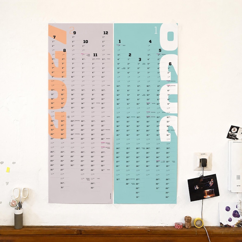 May include: A vertical wall calendar featuring two side-by-side panels. The left panel is light gray with large peach-colored text reading 2023. The right panel is light blue with large white text reading 2024. Both panels display monthly dates.