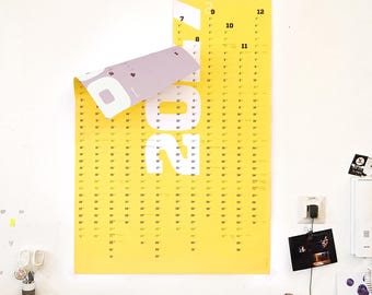 Könnte beinhalten: Ein leuchtend gelber Wandkalender mit dem großen weißen Text 2024. Die obere linke Ecke des Kalenders ist umgeklappt und gibt den Blick auf eine hellviolette Unterseite frei. Der Kalender zeigt Monate und Tage in einem Raster mit schwarzer Schrift.