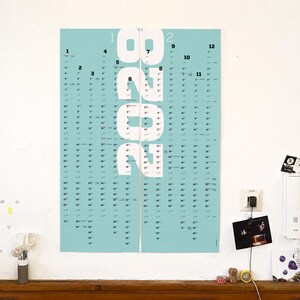 May include: A vertical wall calendar for the year 2028 featuring a light blue background with large white numbers spelling out 2028 in the center. The calendar displays twelve months arranged in columns with black text for dates and month numbers.