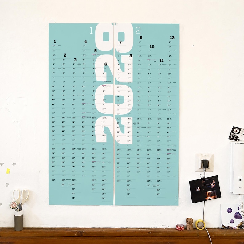 May include: A vertical wall calendar for the year 2028 featuring a light blue background with large white numbers spelling out 2028 in the center. The calendar displays twelve months arranged in columns with black text for dates and month numbers.