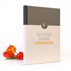 May include: A beige journal with a black spine and a pocket on the front. The journal has the text "SCHÖNE JAHRE CHRONIK" printed on the front in black and orange.