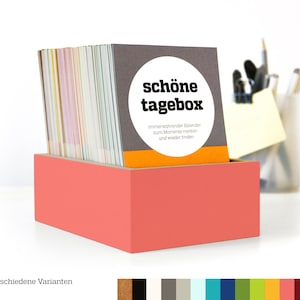 Può includere: Una scatola di colore corallo con un mazzo di carte colorate all'interno. Le carte hanno il testo "schöne tagebox" e "immerwährender Kalender zum Momente merken und wieder finden".