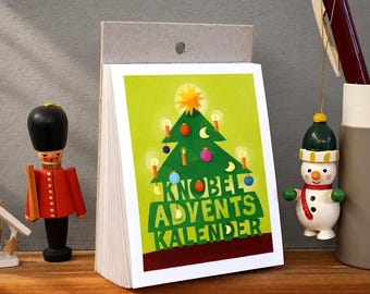 Könnte beinhalten: Ein Papier-Adventskalender mit einer grünen Weihnachtsbaum-Illustration, die bunte Kugeln, Kerzen und einen gelben Stern zeigt. Der Text lautet KNOBEL ADVENTS KALENDER. Er wird neben einer hölzernen Soldatenfigur und einem Schneemann-Anhänger präsentiert.