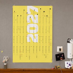 May include: A bright yellow wall calendar for the year 2024 featuring large white vertical text displaying the year. The calendar layout includes twelve columns representing each month, with black numerical dates listed in a vertical grid format.