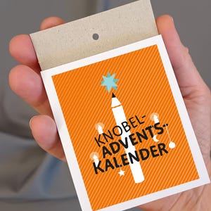 Op de afbeelding: Oranje kaart met een wit potlood in de vorm van een kerstboom met een blauwe ster bovenop. De tekst "Knobel-Advents-Kalender" staat in het zwart op de kaart.