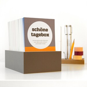 Könnte beinhalten: Eine braune Box mit einem Stapel Karten darin. Die oberste Karte trägt den Text "schöne tagebox" in braunen Buchstaben mit einem weißen Kreis darum. Der Text unter dem Kreis lautet "immerwährender Kalender zum Momente merken und wieder finden."
