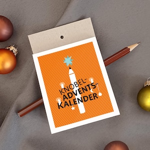 May include: A rectangular card with an orange background featuring diagonal white stripes and a white pencil graphic. The text reads KNOBEL-ADVENTS-KALENDER in black letters. A light blue star sits atop the pencil, surrounded by small white circles and stars. A brown pencil lies diagonally across the card.