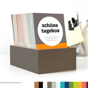 Könnte beinhalten: Eine graue Box mit einem Stapel bunter Karten darin. Die oberste Karte hat einen weißen Kreis mit dem Text "schöne tagebox" in Schwarz. Der Text unter dem Kreis lautet "immerwährender Kalender zum Momente merken und wieder finden".