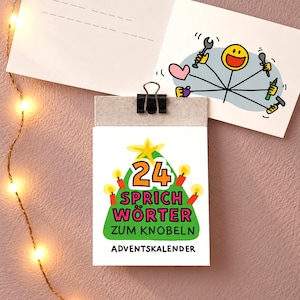 Könnte beinhalten: Ein Abreißkalender mit schwarzer Klemme, verziert mit einem grünen Weihnachtsbaum, einem gelben Stern und vier roten Kerzen. Der Text lautet 24 SPRICH WÖRTER ZUM KNOBELN ADVENTSKALENDER. Darüber liegt eine Karte mit einem gelben Smiley und Werkzeugen.