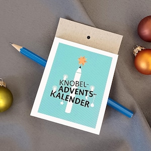 May include: A rectangular card with a light blue background and white diagonal stripes, featuring a white pencil illustration with an orange star and the black text KNOBEL-ADVENTS-KALENDER. A blue pencil rests across the card.