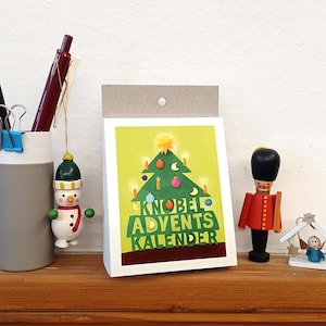 Könnte beinhalten: Eine weiße Karte mit einer grünen Weihnachtsbaum-Illustration und dem Text "Knobel Adventskalender". Die Karte liegt auf einer Holzoberfläche mit einem Schneemann-Anhänger, einem Spielzeugsoldaten und einem kleinen Weihnachtsbaum-Anhänger.