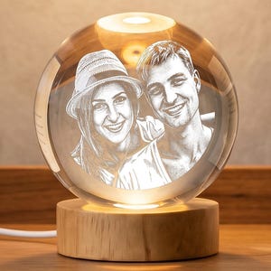 Puede incluir: Un pisapapeles de cristal esférico con un retrato grabado con láser de dos personas, una de ellas con sombrero. La esfera de cristal transparente está iluminada desde abajo por una base circular de madera clara con un cable blanco.