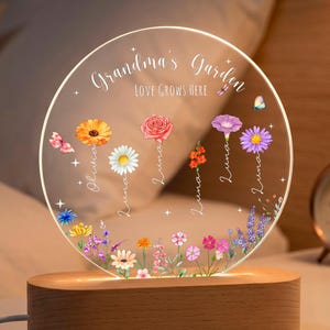 以下が含まれることがあります： 木製ベース付きの円形アクリル製ナイトライト。「Grandma's Garden」および「LOVE GROWS HERE」という文字が印刷されています。様々な花や蝶のカラフルなイラストが描かれ、茎に沿って縦書きで名前が記されています。